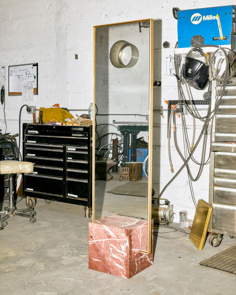 Standing mirror with red jasper cube base and rectangular brass mirror frame in metal workshop.