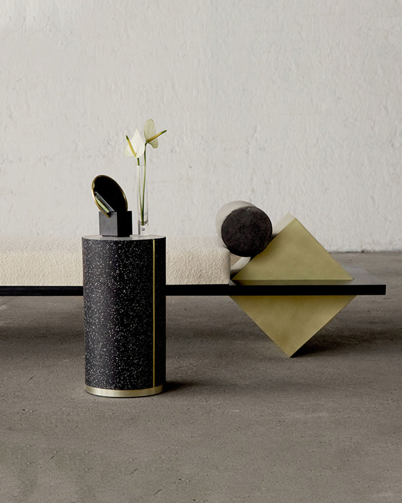 Iimage of daybed showing brass cube base, blackened steel structure. beige boucle mattress and grey velvet bolster pillow. Speckled black rubber CYL side table standing in front of the daybed.