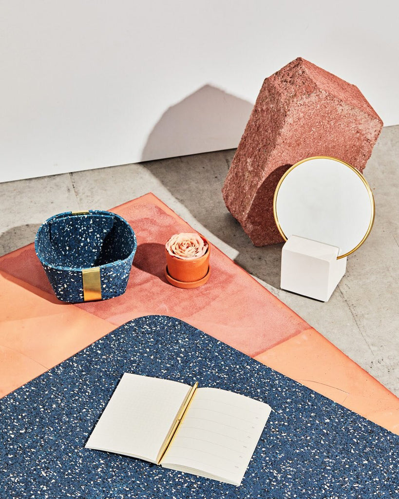 Desk set up with white cube base vanity mirror, small speckled blue rubber & brass basket and  rounded edge speckled blue rubber desk mat with open notebook and brass pen on concrete ground with terracotta surface.