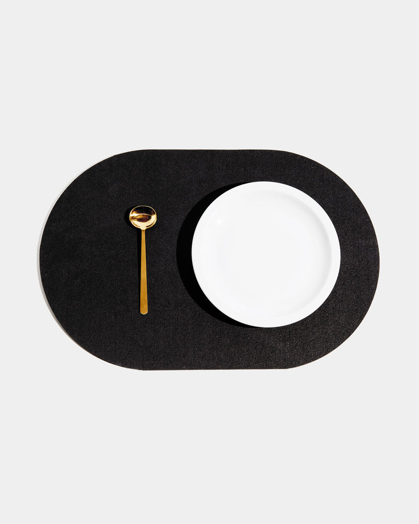 Top view of black rubber capsule placemat with white plate and brass spoon on white background.