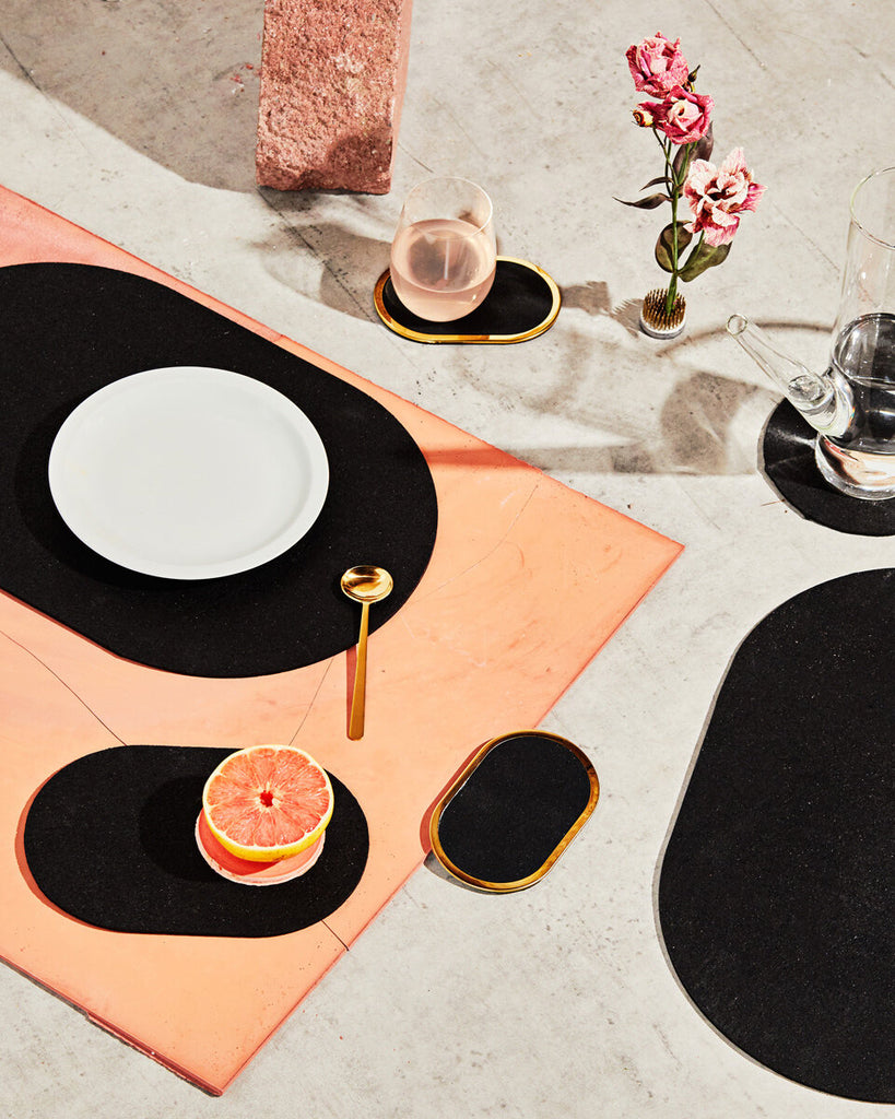 Spring table setting with black rubber capsule placemats, capsule trivets and brass ring coaster. The setting is styled with grapefruit, pink liquid in glass and pink flower.