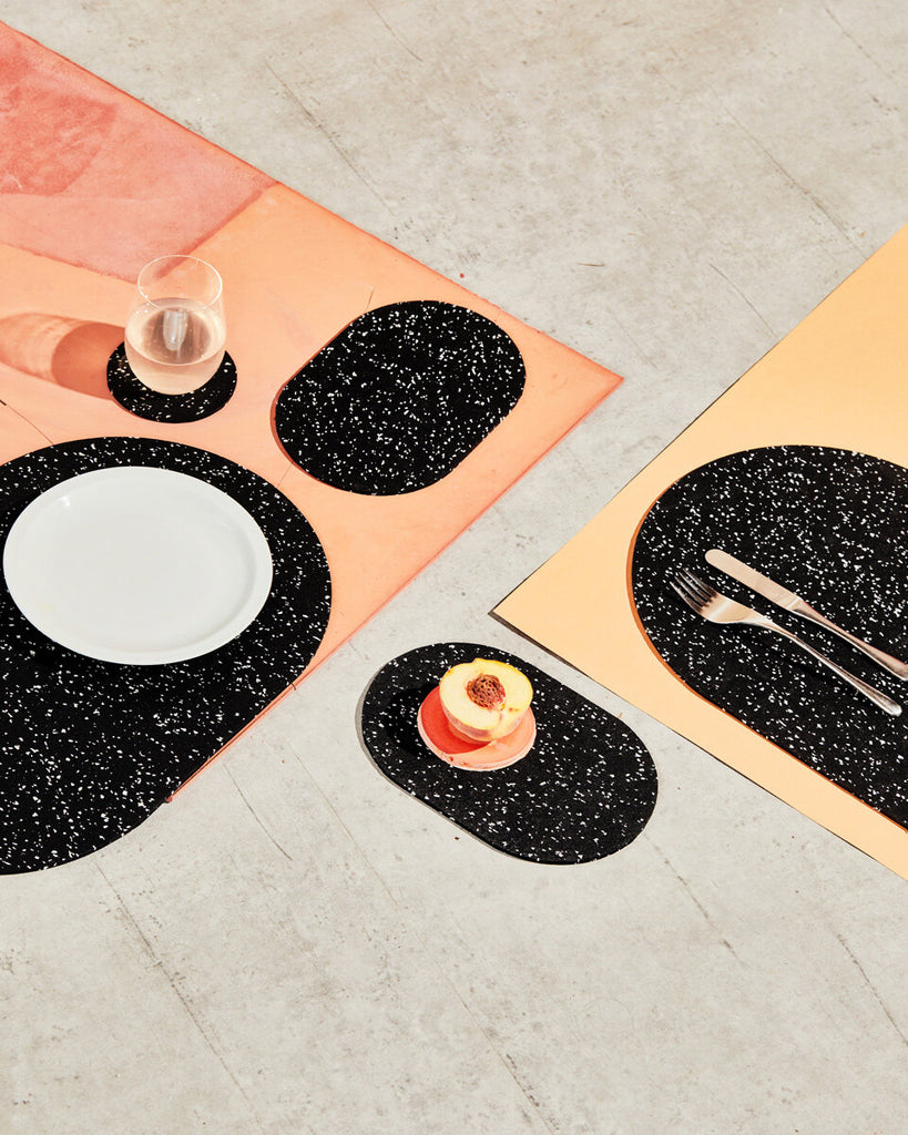 Spring table setting with speckled black rubber capsule placemats and capsule trivets on concrete and terracotta surface. The setting is styled with cut half peach, white plate, glass and cutlery.