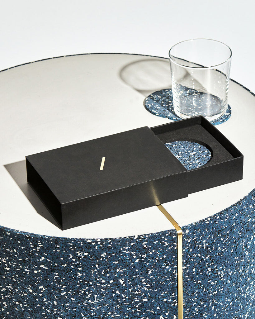 Speckled blue oval rubber coaster inside black rectangular packaging on concrete surface. Oval coaster with empty glass behind the packaging.