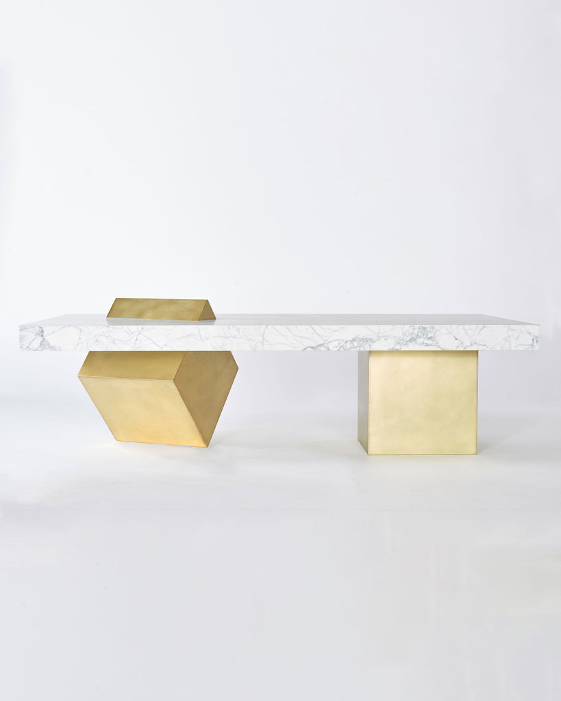 Coffee table with rectangular white carrara marble top and two cube brass legs. One of the brass cube leg is standing on its knife-edge slicing through marble top.