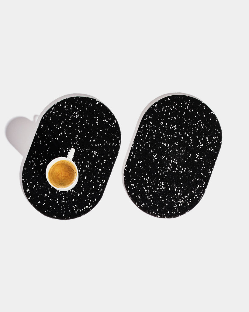 Two capsule speckled black rubber trivets on white surface. One trivet has a white espresso cup on it.